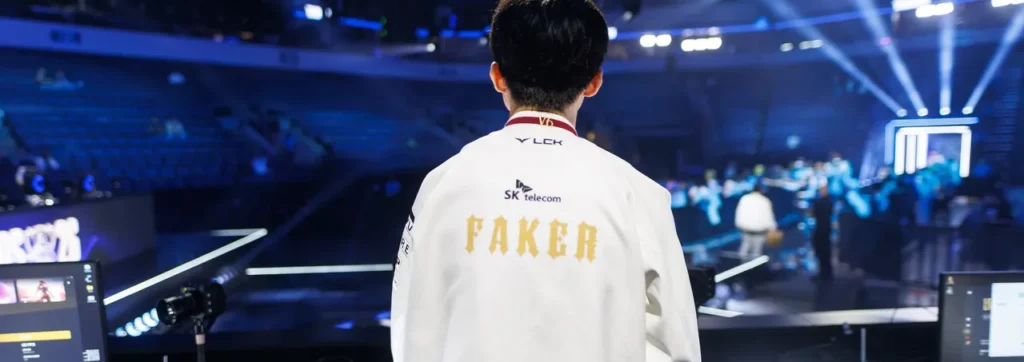 Faker, de costas, com a jaqueta branca da T1 com o nome FAKER em dourado, olha a arena iluminada de azul no palco do Worlds 2026 em Allen