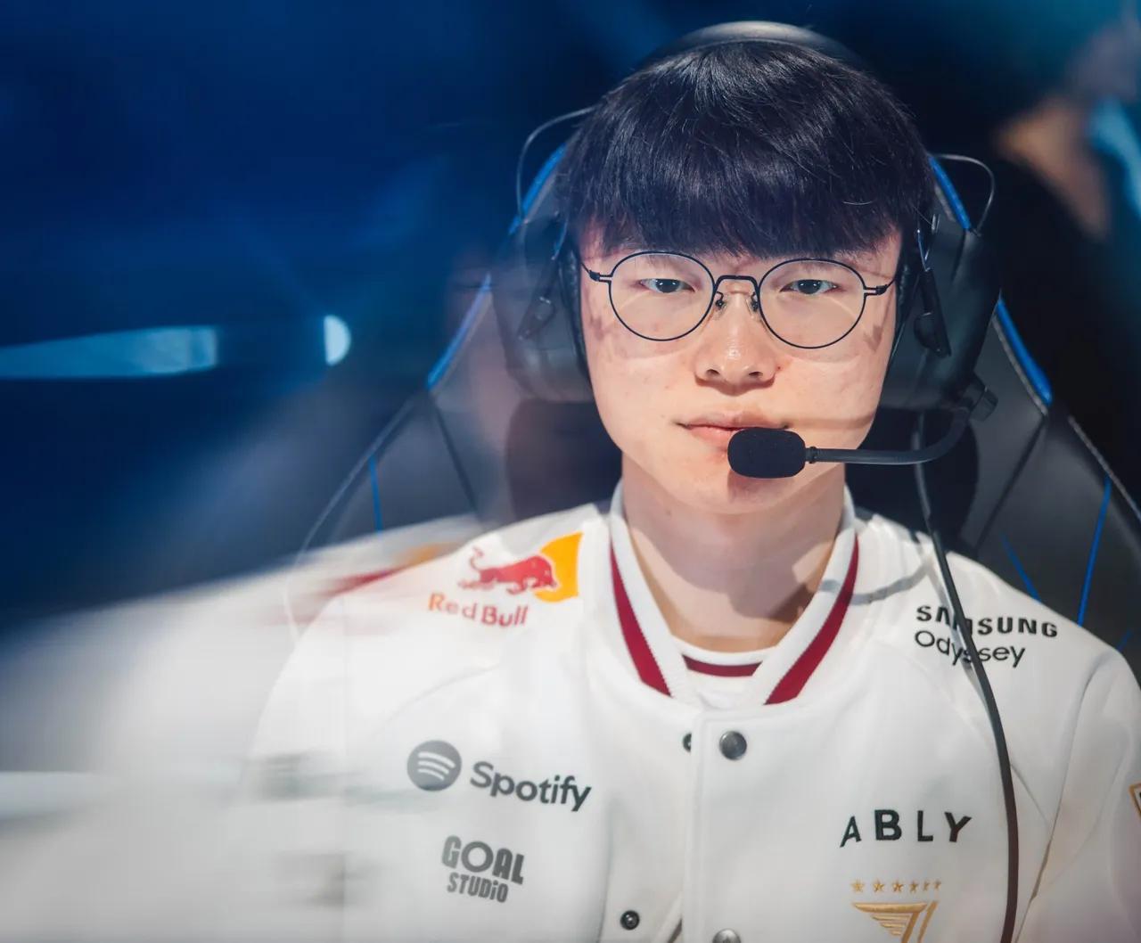 Faker, de óculos e headset, com a jaqueta branca da T1, sentado no palco do Worlds 2026 em Allen