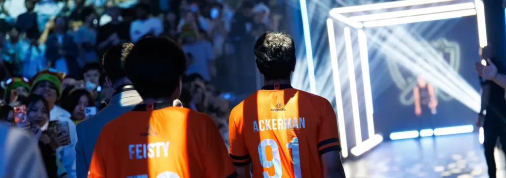 Feisty e Ackerman, da LØS, de costas, com as camisas laranja 70 e 91, caminham diante da torcida para o palco do Worlds 2026 em Allen